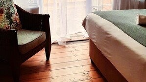 Een kamer met een houten vloer, een bed met een groene spreidekking, een rieten stoel met een bloemenkussen, en dunne gordijnen voor de ramen.