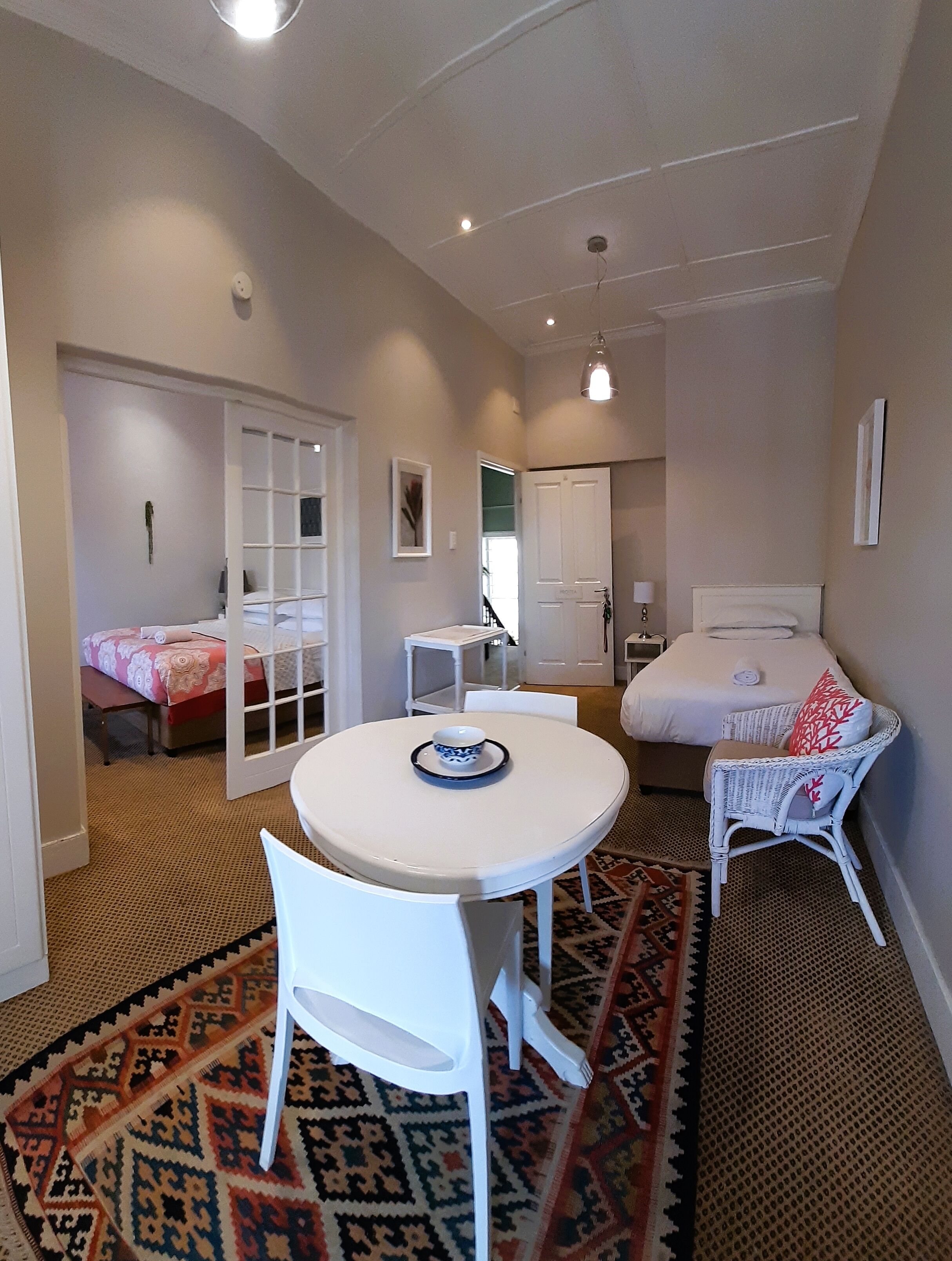 A room with a round white table, a white chair, a bed with a white headboard, and a patterned rug.