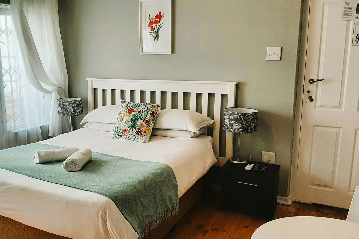 Ein ordentlich gemachtes Bett mit einer grünen Tagesdecke, zwei Kissen und zwei aufgerollten Handtüchern. Darüber hängt ein gerahmtes Blumenbild, ein Nachttisch mit einer Fernbedienung und eine Nachttischlampe.