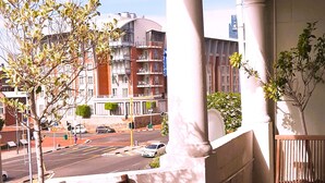 Een balkon met een houten bankje en een kleine tafel, met uitzicht op een straat met gebouwen en een heldere lucht.