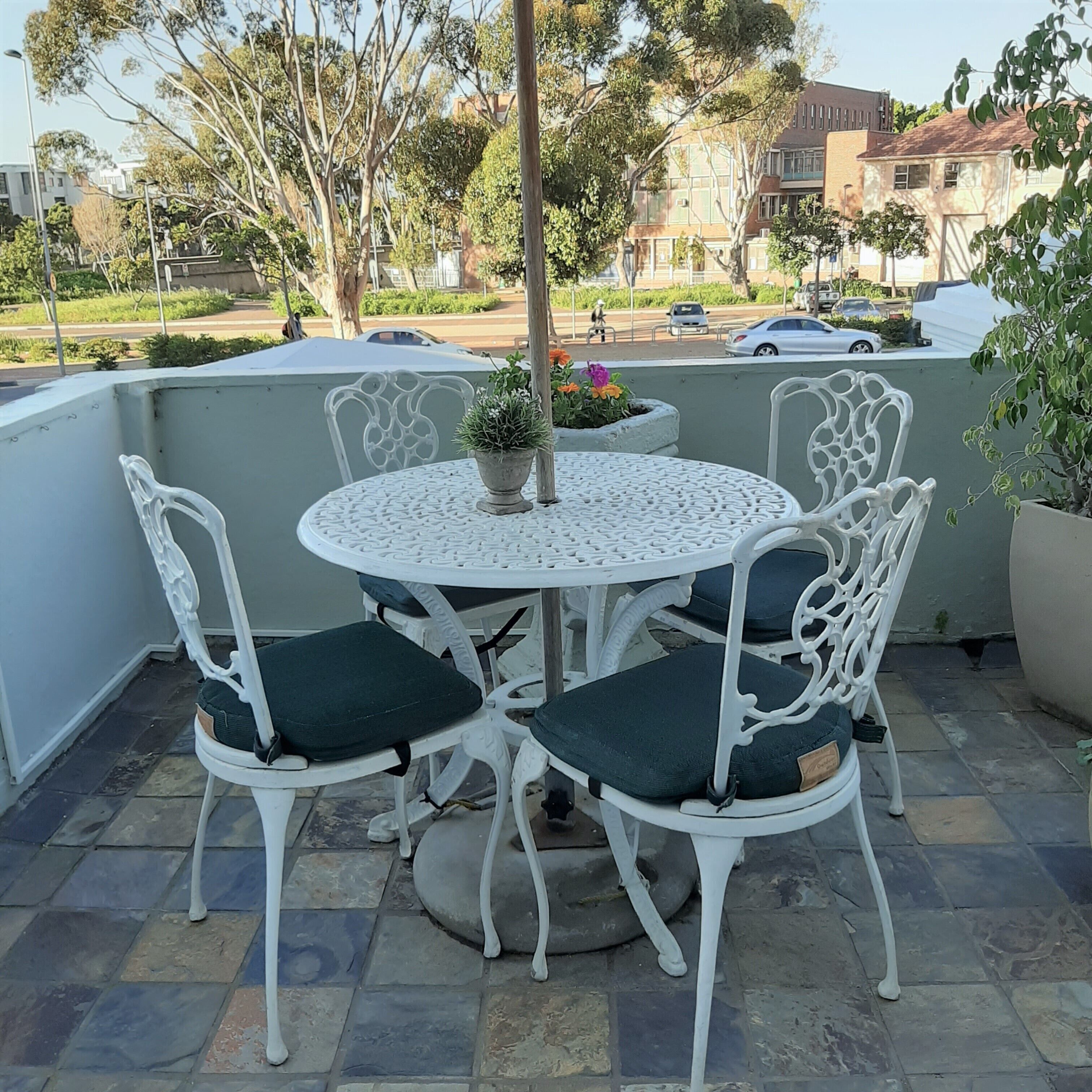 A round outdoor table with white metal chairs and cushions, surrounded by potted plants and a view of the street.