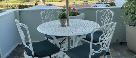A round outdoor table with white metal chairs and cushions, surrounded by potted plants and a view of the street.