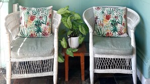 Een kamer met twee rieten stoelen, een bijzettafeltje met een plant, een wandklok, een kaart en hangplanten.