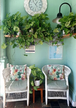 A room with two wicker chairs, a small table with a plant, a wall clock, a map, and hanging plants.