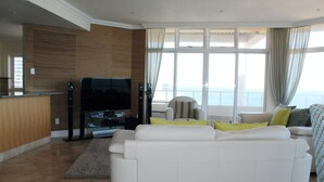 A modern living room with a white sofa, a flat-screen TV, and a view of the sea through large windows. - Unit 1101 Oyster Rock (Umhlanga)