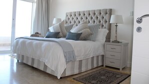 A bedroom with a tufted headboard, a bed with striped bedding, two bedside lamps, a nightstand with drawers, and a patterned rug. - Unit 1101 Oyster Rock (Umhlanga)