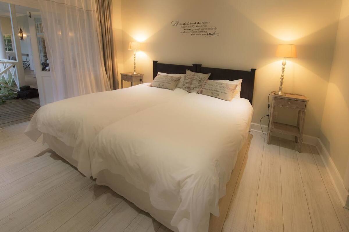 A large bed with white linens and multiple pillows, two bedside lamps, and a wall quote.