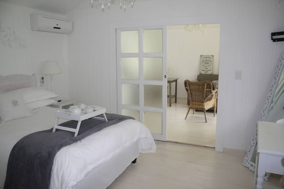 A bedroom with a bed, bedside table, and a tray with tea set. There is a door leading to a dining area with a chair and a table.