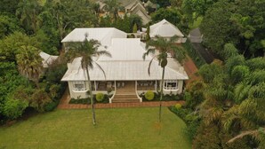 A large, white, two-story house with a covered porch and a small balcony, surrounded by lush greenery and palm trees. - Lemon Tree B&B (Kloof)