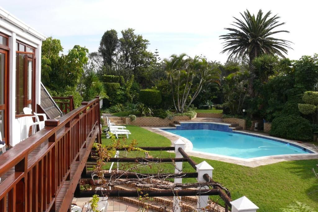 A swimming pool surrounded by lush greenery, a wooden deck with chairs, and a white building with large windows.