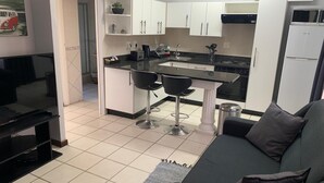 A modern kitchen with white cabinets, a black countertop, and a central island with two bar stools. There is a refrigerator, a sink, and a stove. - Aberdour Guesthouse (Gqeberha (Port Elizabeth))