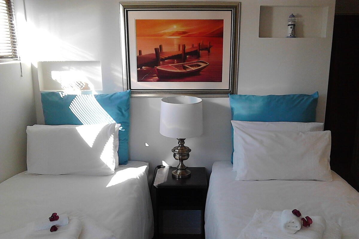 A hotel room with two beds, a nightstand with a lamp, a painting of a sunset over a harbor, and a small shelf with a decorative item.