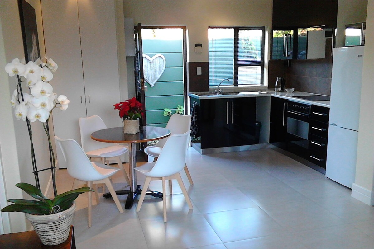A modern kitchen with white cabinets, a black countertop, and a white refrigerator. There is a dining area with a round table and white chairs.