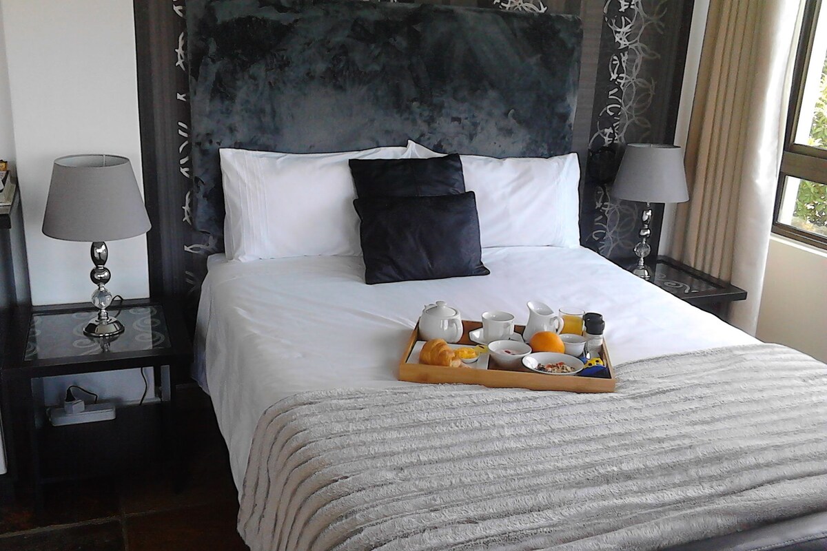 A neatly made bed with a tray of breakfast items, a bedside table with a lamp, and a window with curtains.