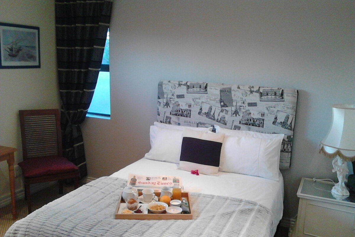 A hotel room with a bed, a tray of breakfast items, a chair, a nightstand, and a window with curtains.