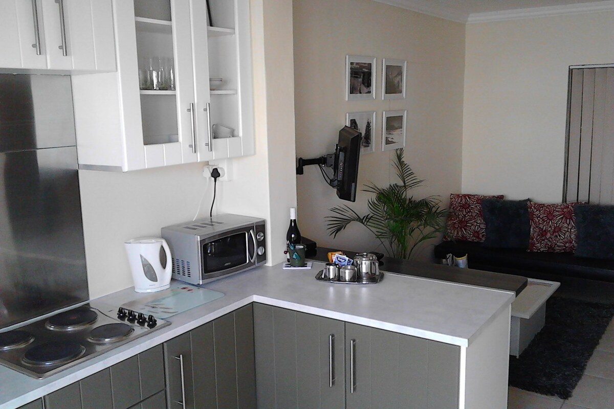 A compact kitchen with a microwave, kettle, and stove. There is a dining area with a sofa and a plant.