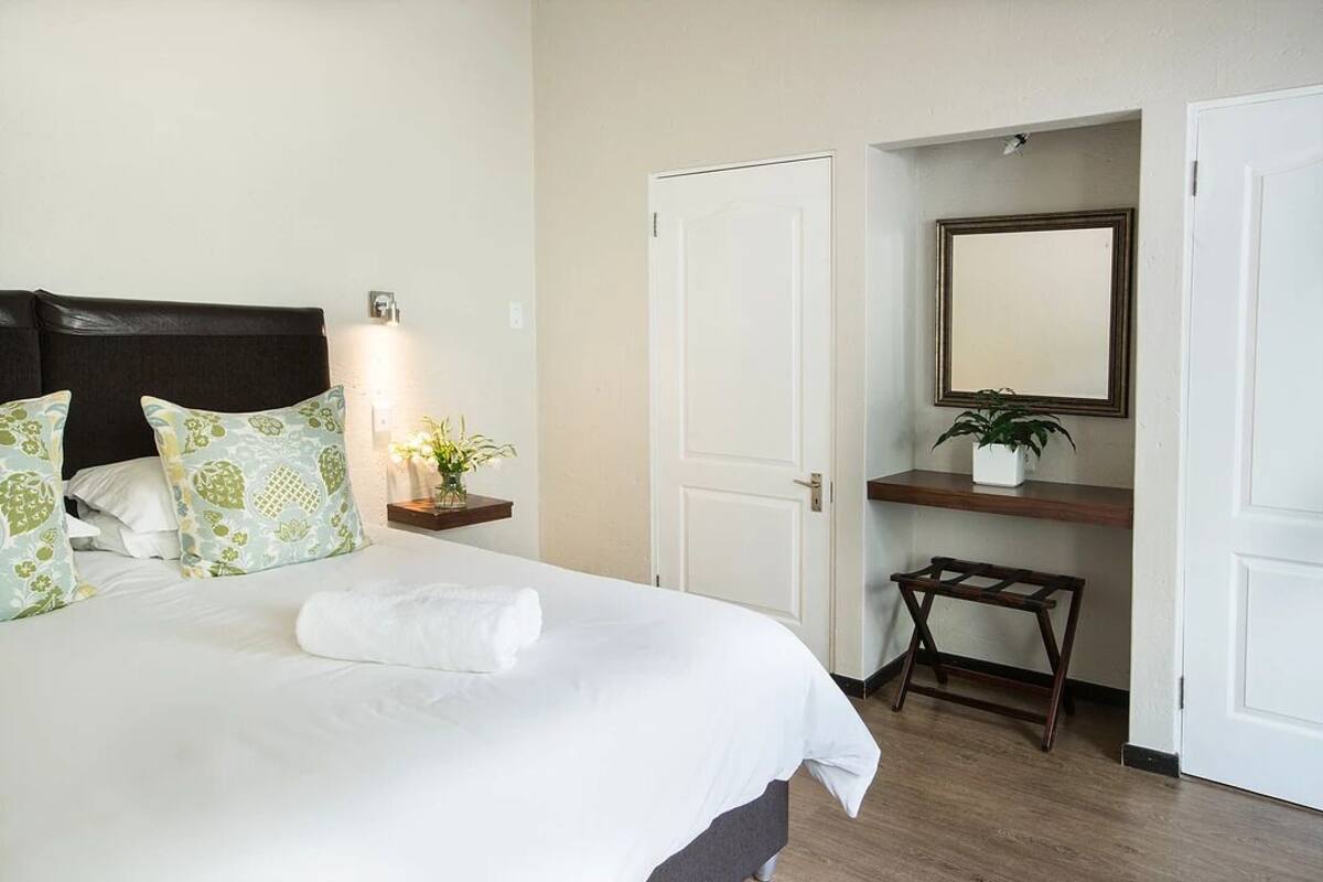 A neatly made bed with a white comforter and green patterned pillows, a bedside table with a plant, a mirror above a wooden shelf, and a small stool.