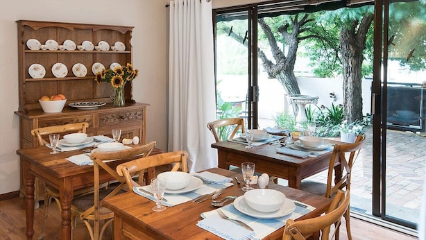 A dining area with a wooden table set for four, surrounded by chairs. A wooden cabinet with dishes is to the left. - Sandton Slippers (Sandton)