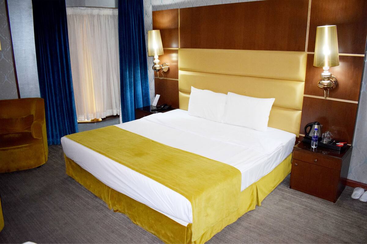 A hotel room with a large bed, a wooden headboard, two bedside lamps, a nightstand, a chair, and a window with curtains.