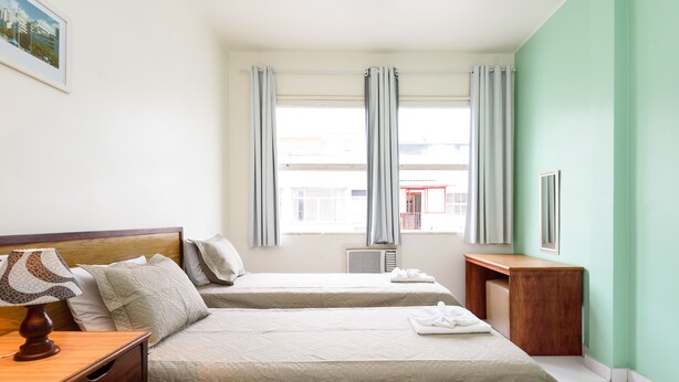 A hotel room with two beds, a wooden nightstand, a lamp, a framed picture on the wall, and a window with curtains. - Omar do Rio - A1208 (Rio de Janeiro)