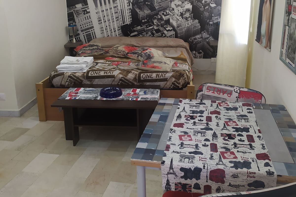 A bedroom with a bed, a desk, and a large cityscape tapestry on the wall.