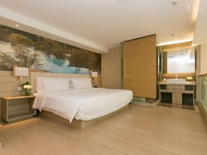 A modern hotel room with a large bed, a nightstand with a vase of flowers, a painting on the wall, and a bathroom with a mirror and sink.