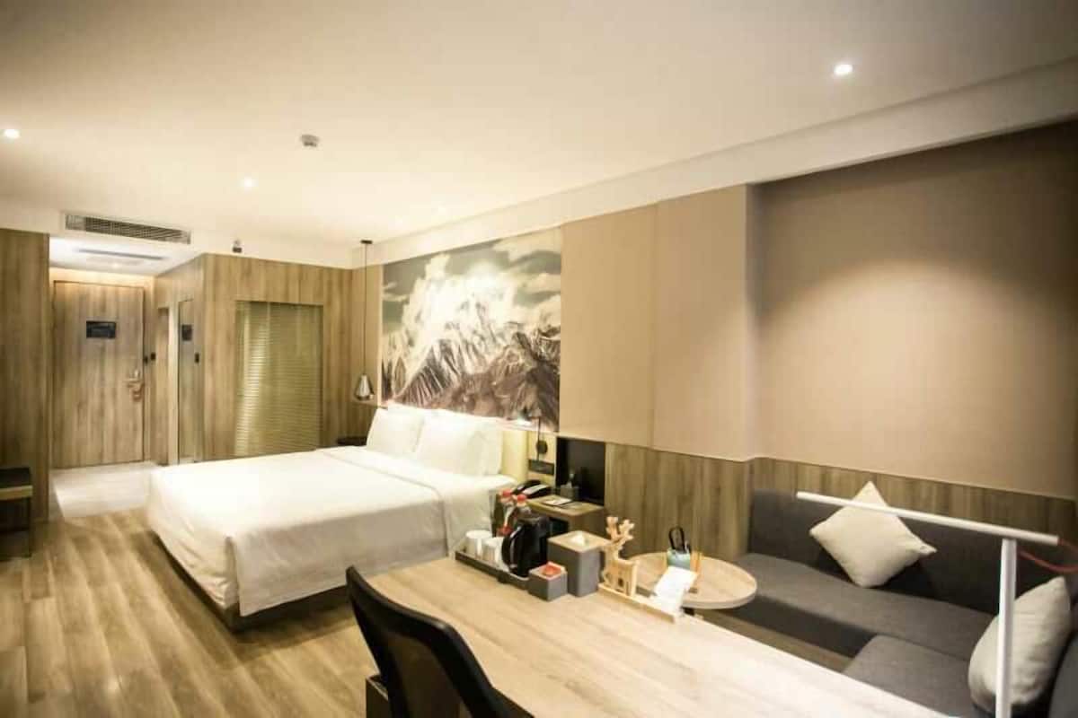 A hotel room with a bed, a desk with a lamp, a sofa, and a wall with a mountain painting.
