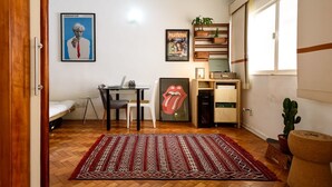 A compact living space with a bed, a desk with a laptop, a framed poster, a bookshelf, and a red patterned rug. - Omar do Rio - BR902 (Rio de Janeiro)