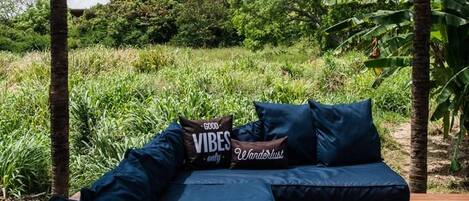 A wooden deck with a sofa and a daybed, surrounded by lush greenery and palm trees.
