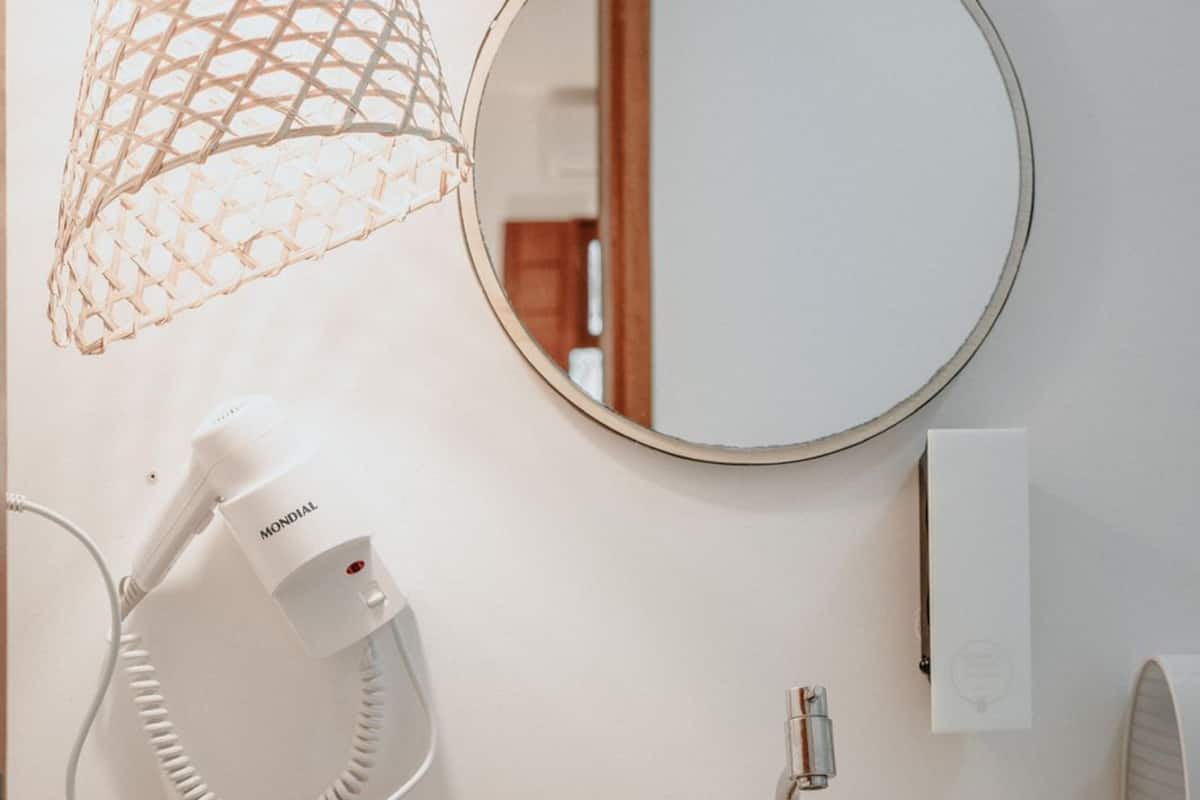 A bathroom with a white sink, a round mirror, a hairdryer, and a pendant light with a woven design.