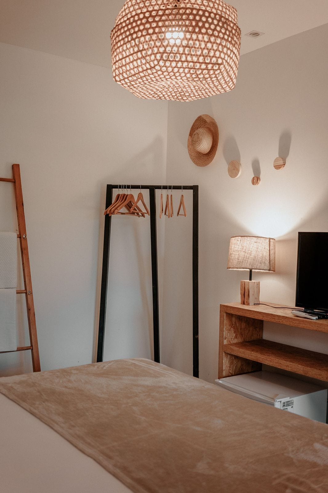 Een slaapkamer met een bed, een houten bureau, een televisie, een hanglamp en een kapstok met hangers.