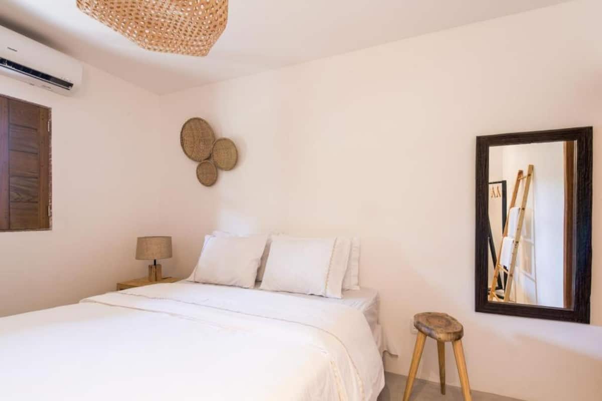 A bedroom with a bed, a nightstand, a lamp, a small stool, a woven wall hanging, and a mirror with a ladder.