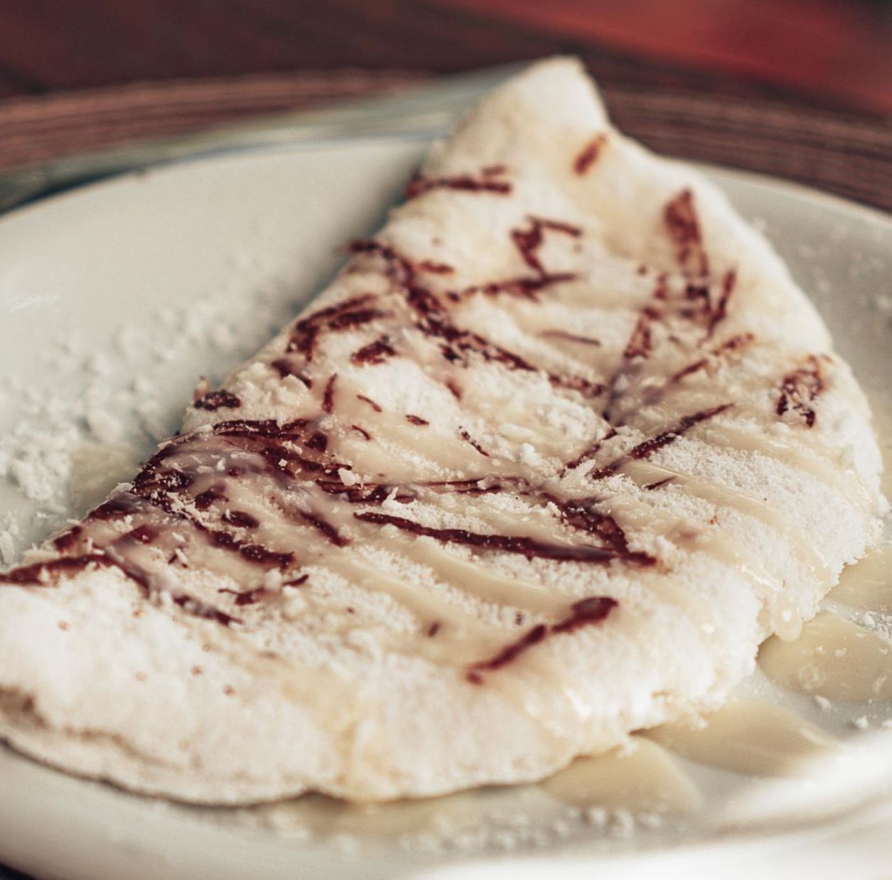 A plate with a piece of food that appears to be a crepe or pancake, lightly dusted with powdered sugar and possibly drizzled with chocolate.