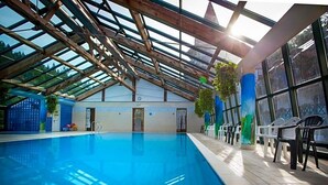 An indoor swimming pool with a glass ceiling, wooden beams, and a row of plastic chairs along the side.
