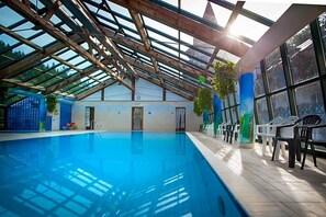 An indoor swimming pool with a glass ceiling, wooden beams, and a row of plastic chairs along the side.