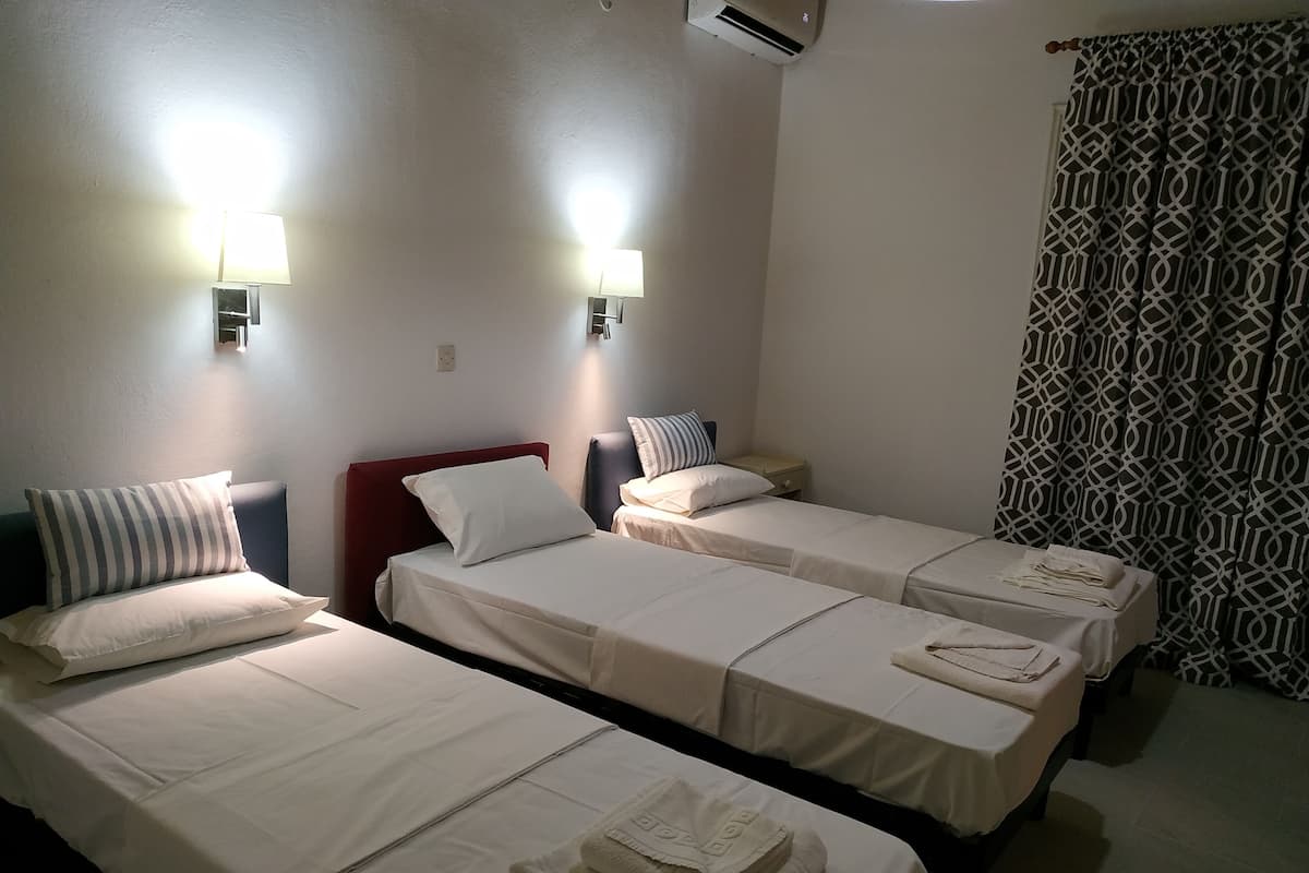 A hotel room with three single beds, each with a white bedspread and pillows. There are two wall-mounted lamps on either side of the beds. The room has a patterned curtain on one side and a plain wall on the other.