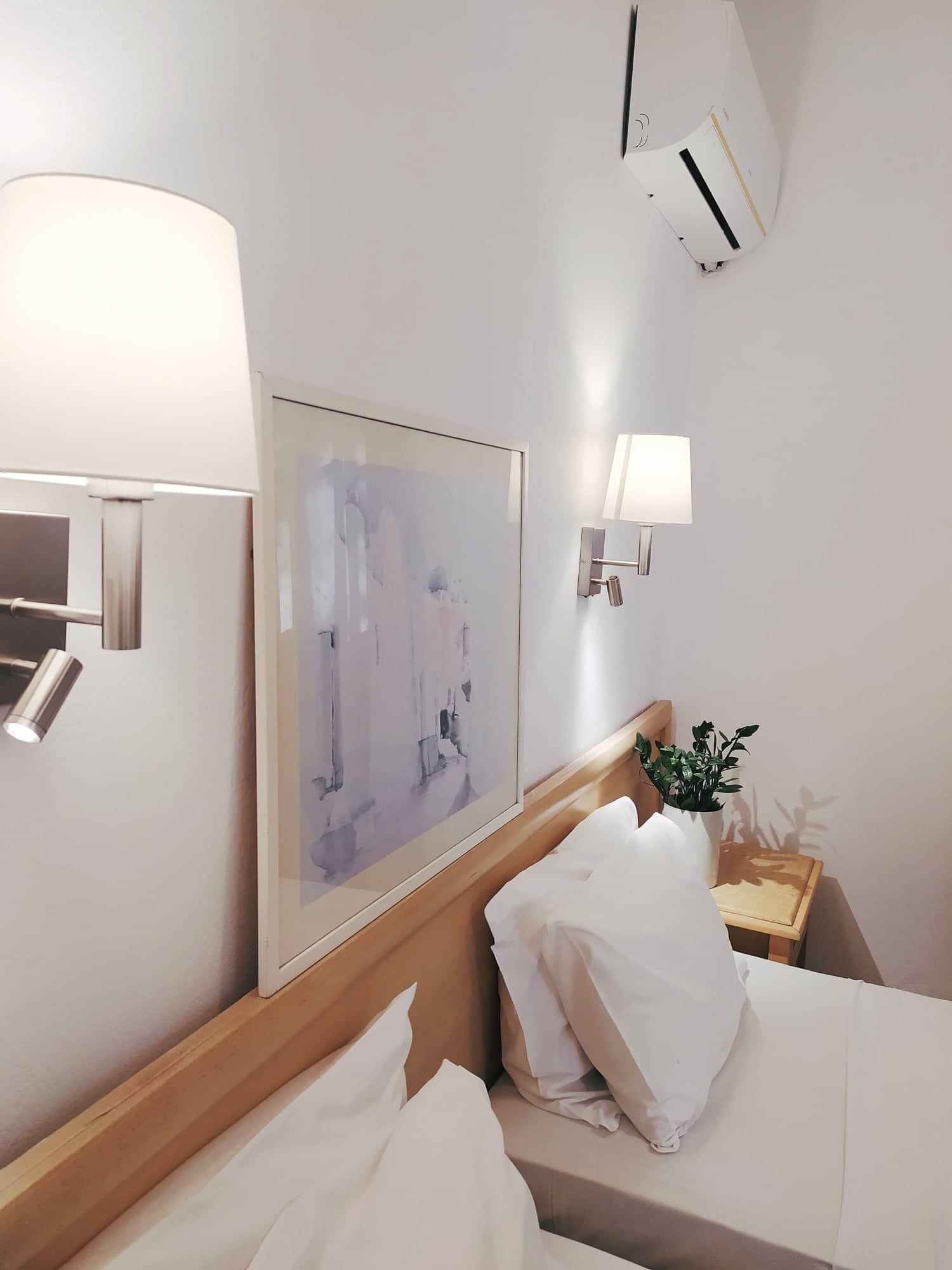 Habitación de hotel con cama, mesita de noche con planta, cuadro abstracto enmarco y luces fijadas a la pared.