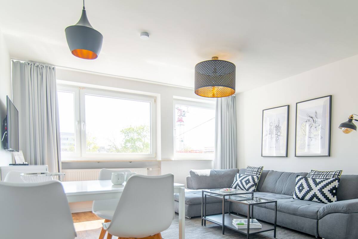 Un salon moderne avec un canapé gris, des fauteuils blancs, une table basse et une grande fenêtre avec des rideaux.