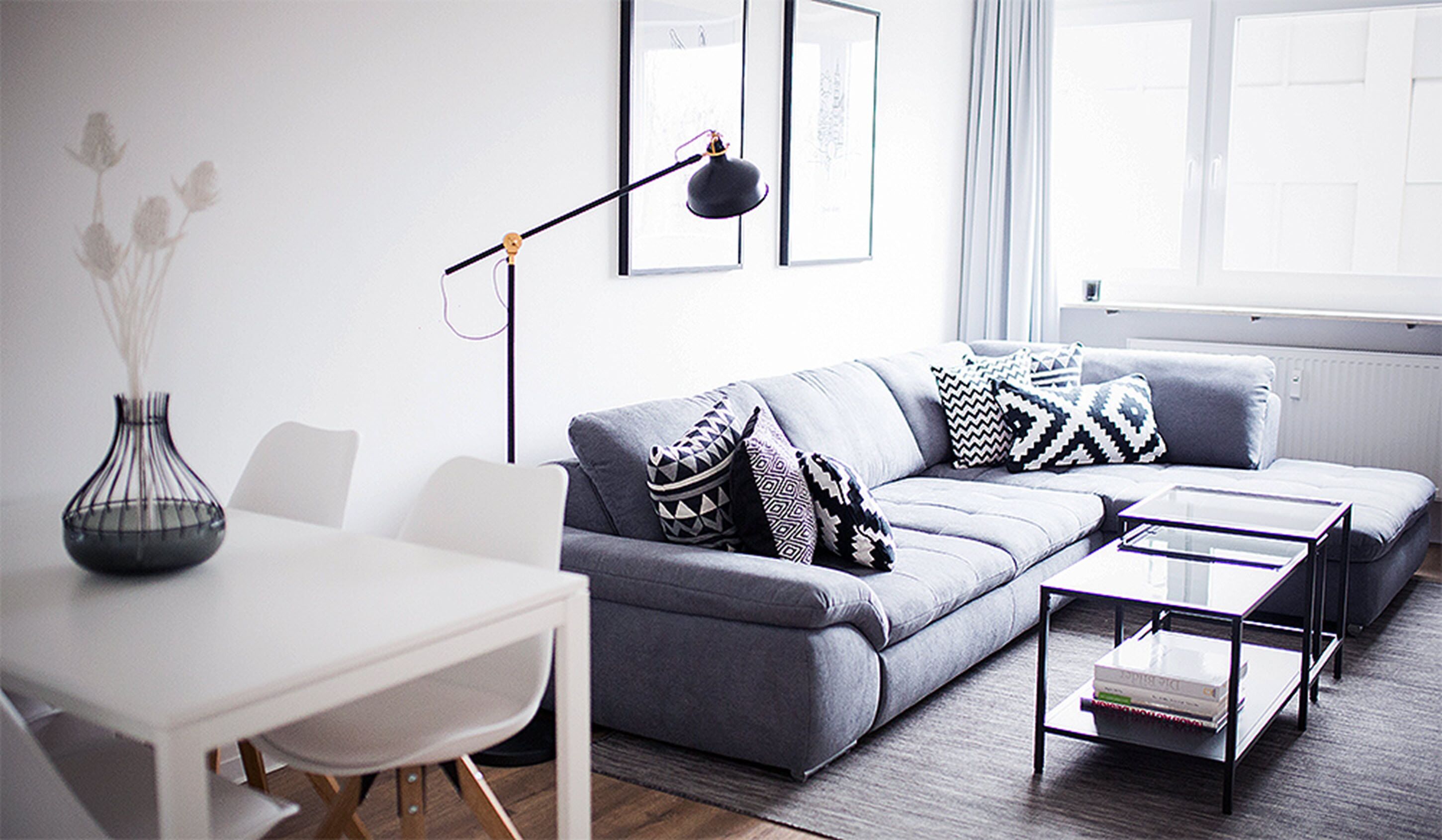 A modern living room with a grey sofa, patterned cushions, a white dining table, and a floor lamp.
