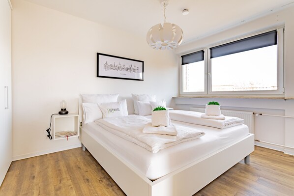 A modern bedroom with a large bed, a bedside table with a lamp, a framed cityscape picture, and a window with blinds.