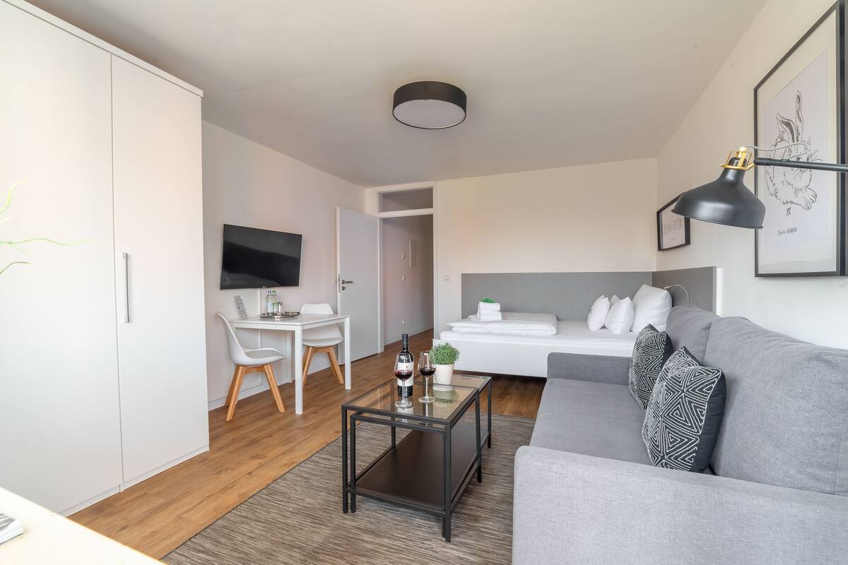 Un salon moderne avec un canapé gris, une table basse en verre, un téléviseur et un lit avec des coussins.