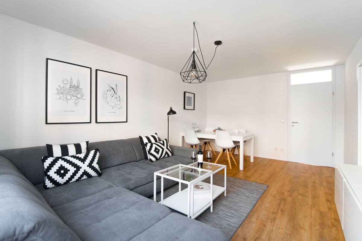 Un salon moderne avec un canapé gris, une table basse en verre et un coin repas comprenant une table et des chaises blanches.