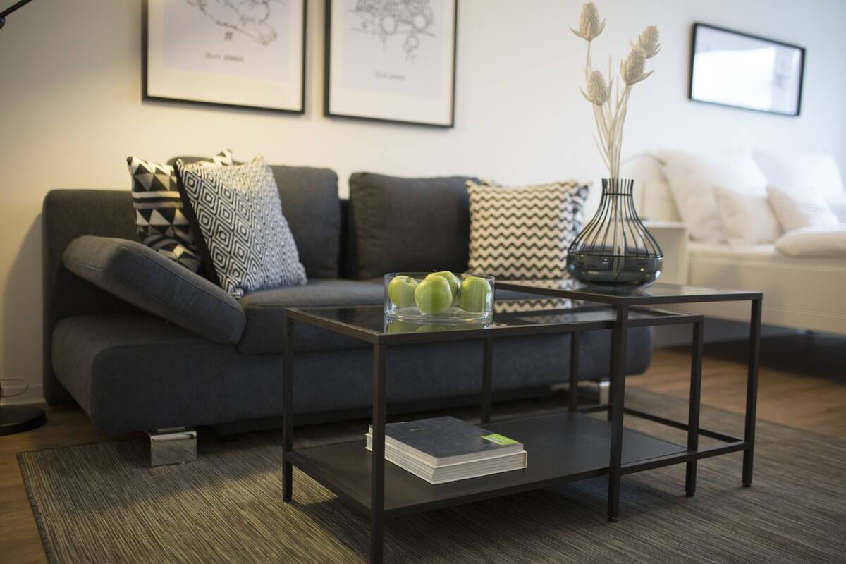Un salon moderne avec un canapé gris, des coussins à motifs, une table basse en verre sur laquelle reposent un livre et un vase de fleurs, et des œuvres d’art encadrées accrochées au mur.