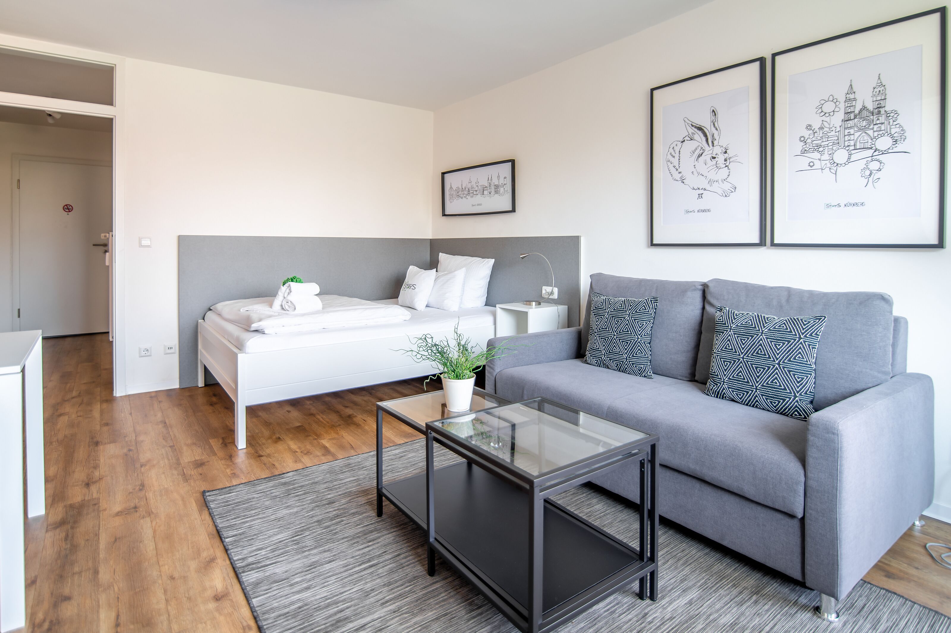 Un dormitorio moderno con una cama, un sofá gris, una mesa de centro de vidrio y dos cuadros en la pared.