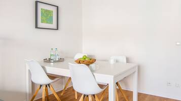 A modern dining area with a white table, four white chairs with wooden legs, a framed picture on the wall, and a bowl of green apples on the table.