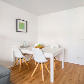 A modern dining area with a white table, four white chairs with wooden legs, a framed picture on the wall, and a bowl of green apples on the table.