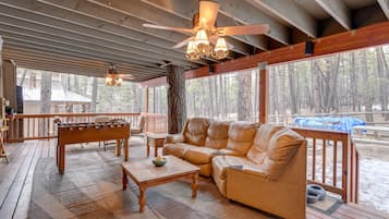 A covered outdoor area with a wooden deck, a leather sofa, a wooden coffee table, and a foosball table.