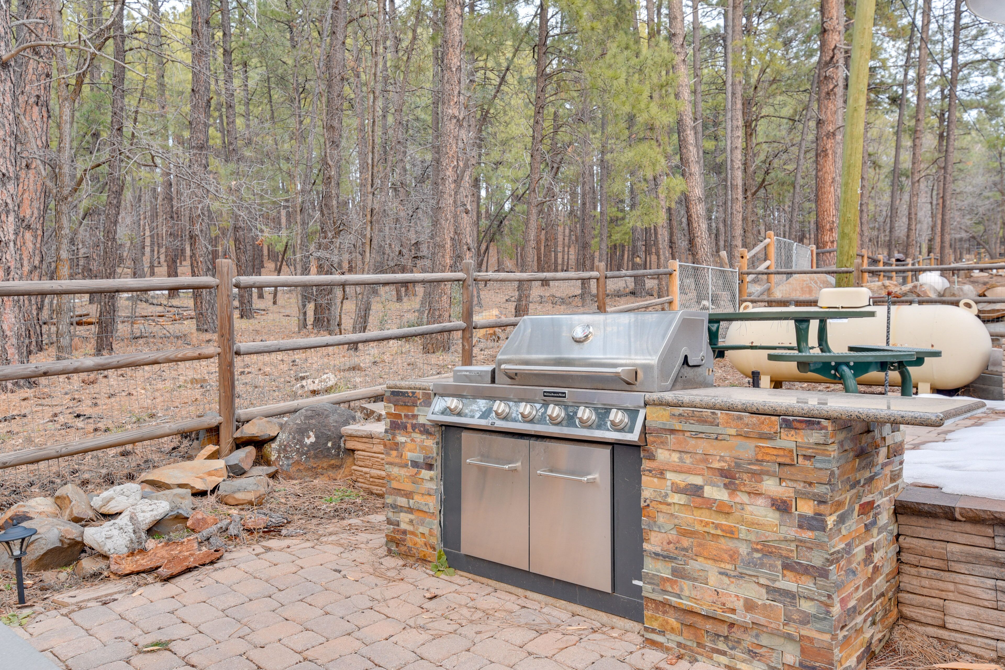 Un espace barbecue extérieur doté d’un barbecue en pierre et en métal, entouré d’une clôture en bois et agrémenté d’une table de pique-nique.