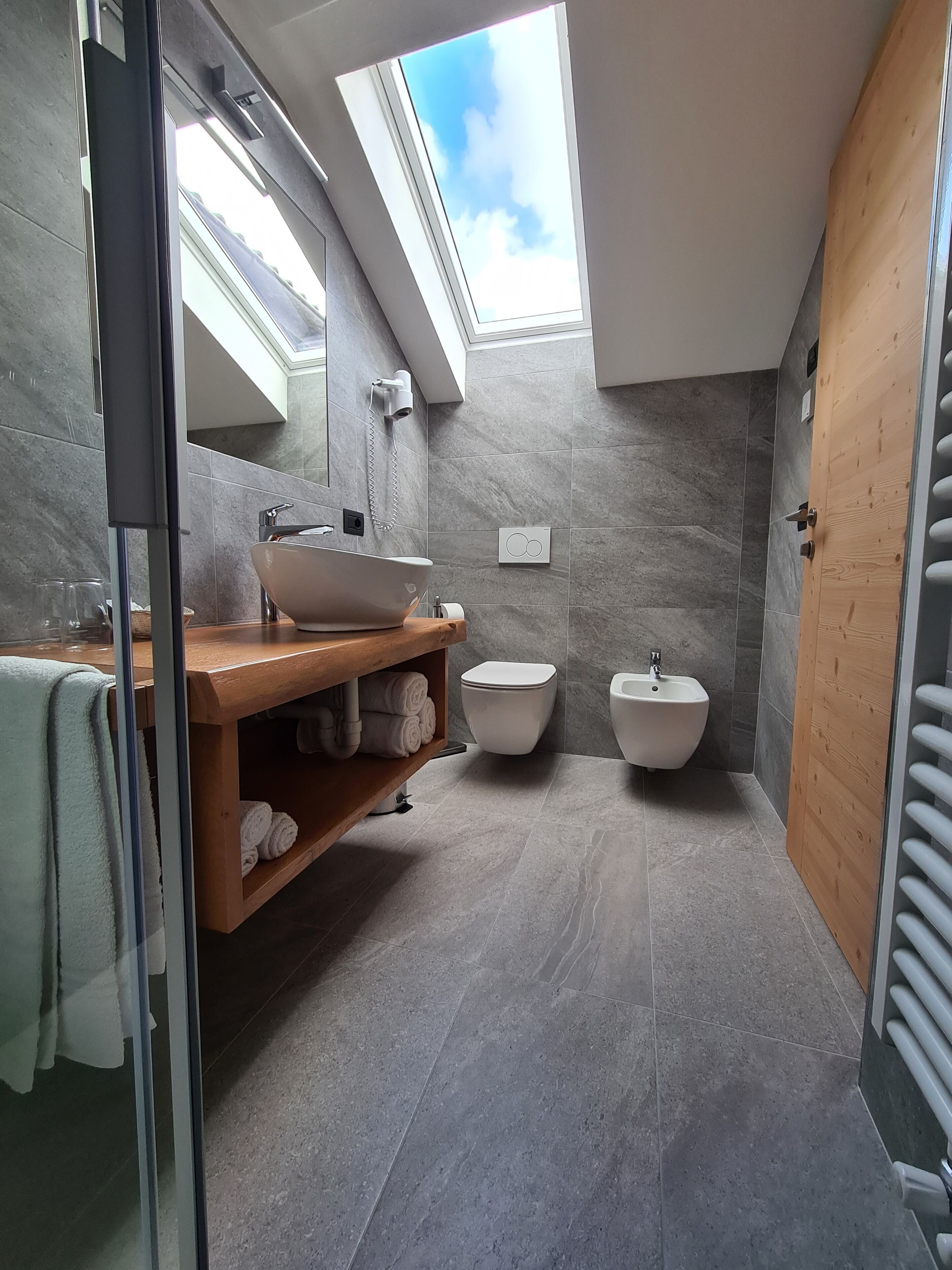 A modern bathroom with a skylight, a wooden vanity, a sink, two bidets, and a towel rack.