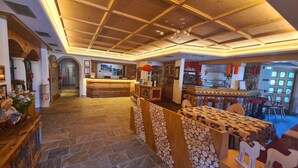 A hotel lobby with a wooden reception desk, a bar area, and a stone-tiled floor. - Hotel Piedibosco (Moena)